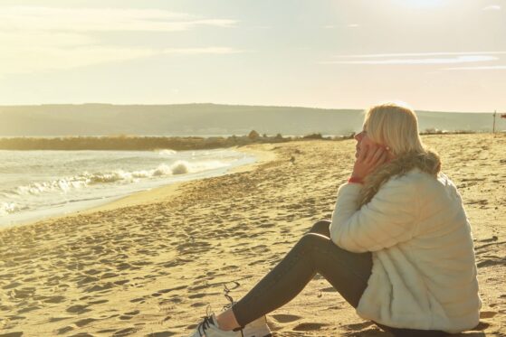 middle-aged-woman-with-blonde-hair-sits-sandy-beach-looking-out-sea